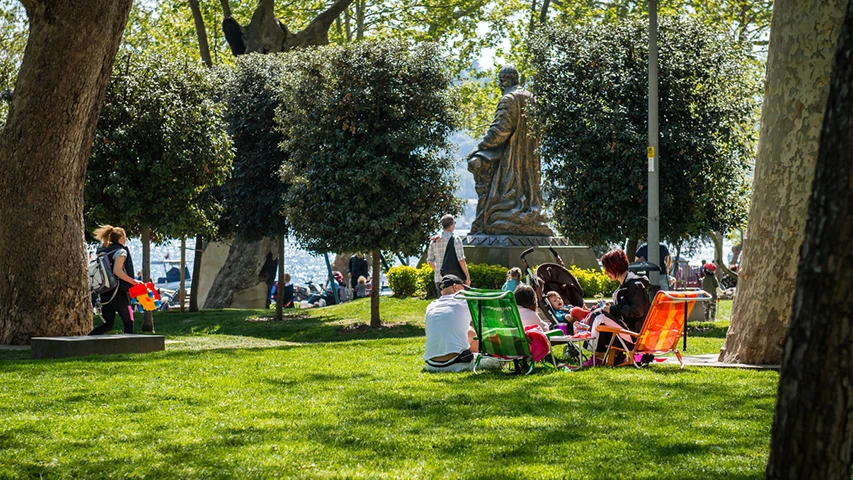 Güneşli bir günde, İstanbul'un Bebek sahilindeki yemyeşil parkta, büyük ağaçların gölgesinde oturan aileler ve bebek arabalarıyla gezinen insanlar, arka plandaki masmavi deniz ve tarihi heykel manzarasıyla huzurlu bir atmosfer sunuyor.