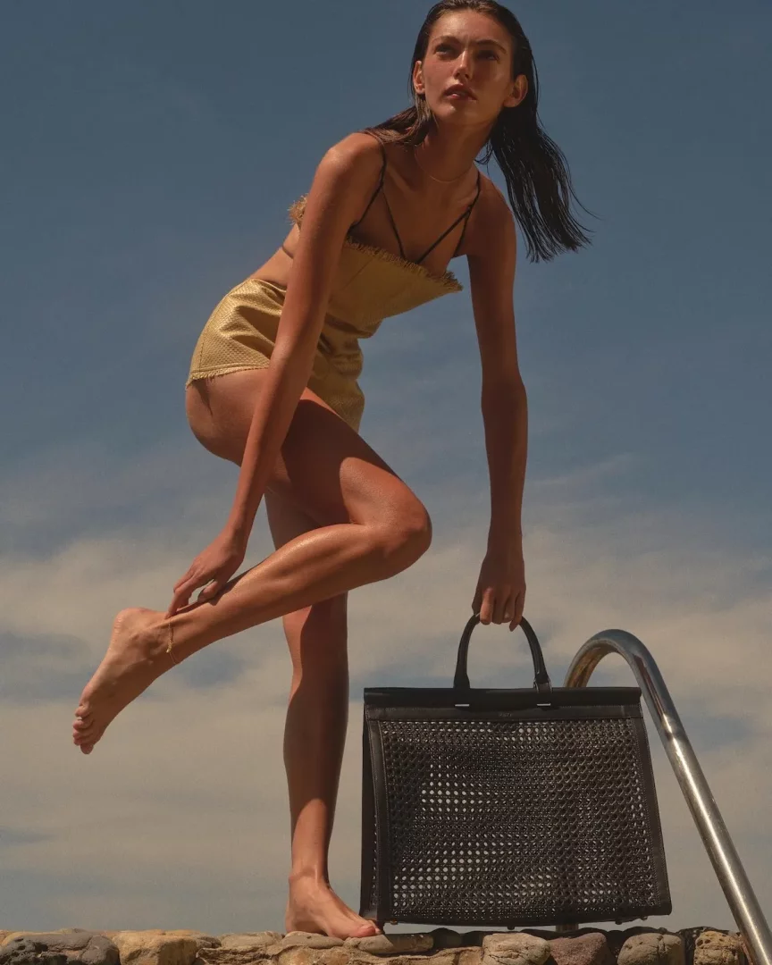 Güneşli bir günde, havuz kenarındaki kayalıkların üzerinde duran, ıslak saçları ve altın sarısı püsküllü bikini takımıyla poz veren genç bir kadın, elinde modern tasarımlı, delikli siyah bir yaz çantası tutarak yukarı doğru bakıyor.