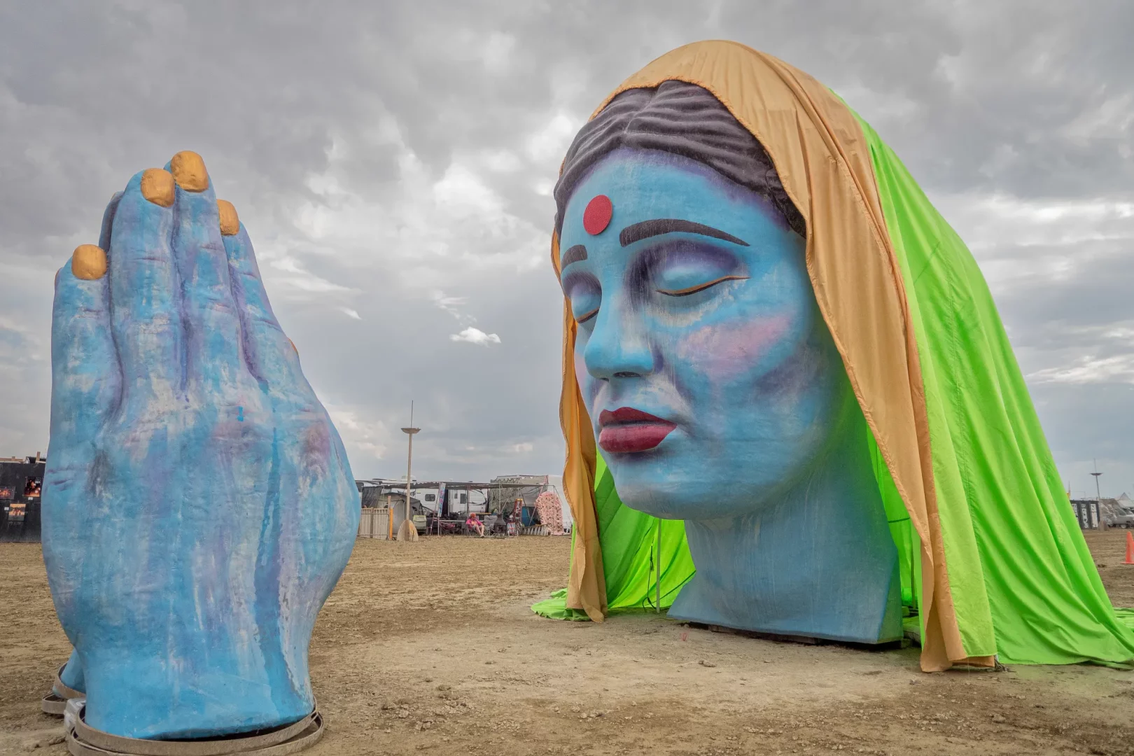Bulutlu bir gökyüzünün altında, Burning Man etkinliğinden bir görselde, açık alanda çöl zemini üzerinde duran, gözleri kapalı, kırmızı bindi ve dudakları olan, sarı ve neon yeşili kumaşlarla örtülü devasa mavi bir kadın başı heykeli ile yanında dua eden bir el heykeli dikkat çekiyor.