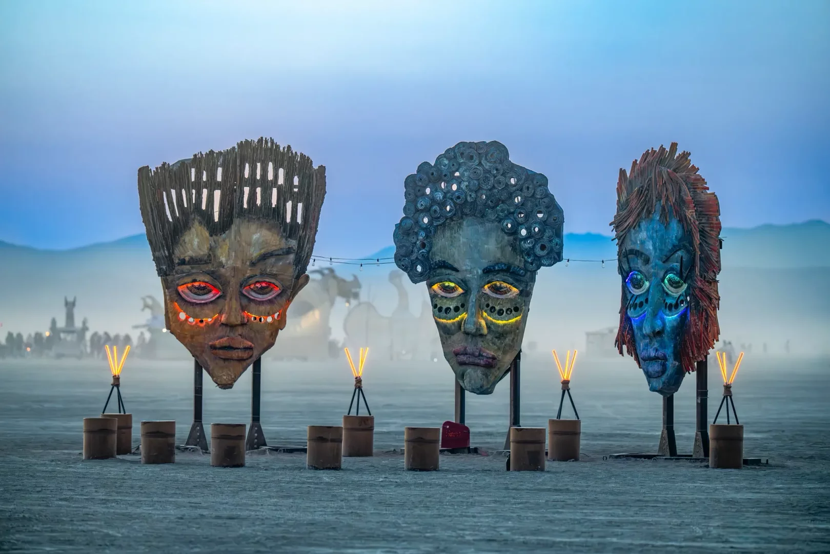 Burning Man festivali alanında, çöl zemini üzerinde sıralanmış, her biri özgün dokulara ve saç şekillerine sahip, gözlerinden ve yüz hatlarından sırasıyla canlı kırmızı, sarı ve mavi ışıklar saçan üç büyük ve dikkat çekici maske heykeli, alacakaranlıkta diğer sanat enstalasyonları ve kalabalıkla çevrili bir manzara sunuyor.