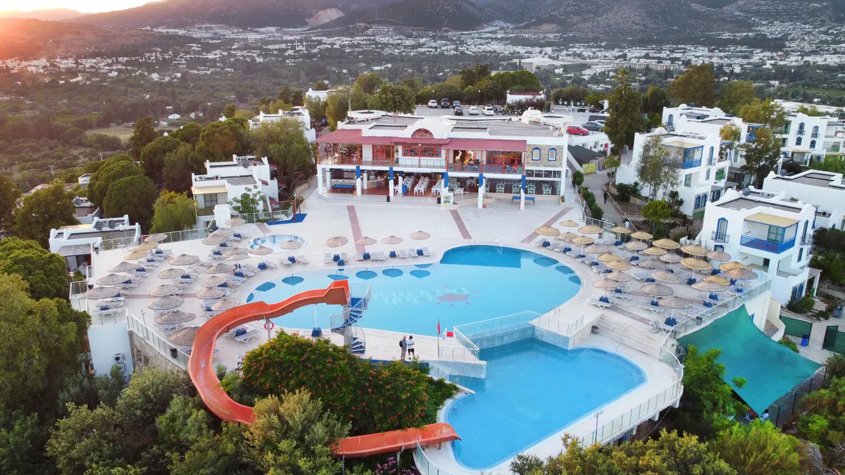 Club Dedeman Bodrum tatil köyünün kuşbakışı görüntüsünde, büyük ana havuzu, spiral su kaydırağı, yemyeşil bitki örtüsüyle çevrili beyaz binaları ve arkada uzanan tepelik bir şehir manzarasıyla keyifli bir konaklama ortamı gözler önüne seriliyor.