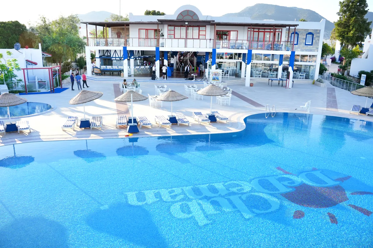 Club Dedeman Bodrum havuzunun dibinde Club Dedeman yazısı ve güneş logosu belirgin bir şekilde görünen, çevresinde şezlonglar, hasır şemsiyeler ve dinlenen insanlar bulunan, arkasında ise tesis binası ve yeşil tepelerin uzandığı geniş ve masmavi bir havuz manzarası.