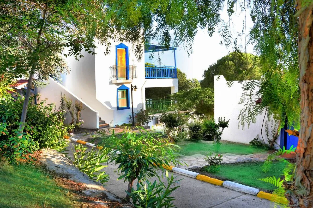 Beyaz duvarlı, mavi pencereleri ve balkonlarıyla Bodrum mimarisine sahip bir tatil köyü binası, yemyeşil ağaçlar ve çimenlerle çevrili bir bahçe yolunun kenarında güneşli bir havada görülüyor.