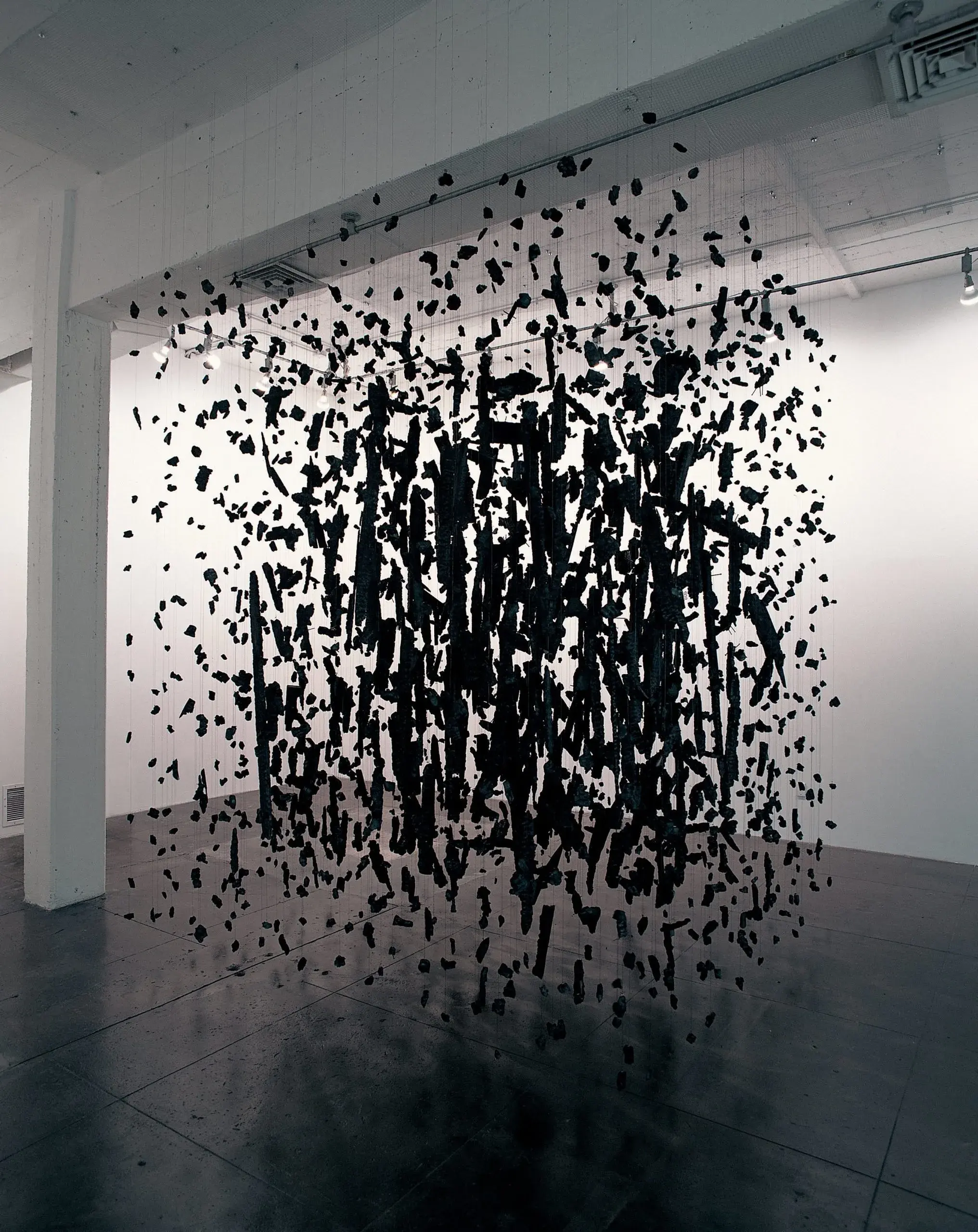 Cornelia Parker'ın Cold Dark Matter: An Exploded View enstalasyonunda, beyaz duvarlı bir galeri boşluğunda, tavandan ince iplerle asılmış yüzlerce kömürleşmiş ahşap parçasının bir patlamanın donmuş anı gibi havada asılı durduğu, dramatik ve etkileyici bir sanat eseri.