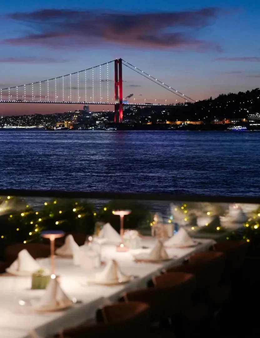 Boğaz'ın üzerinde alacakaranlıkta beyaz ve kırmızı ışıklarıyla parlayan Boğaziçi Köprüsü ve büyüleyici İstanbul silüetinin eşlik ettiği, beyaz örtülü masaların zarif lambalarla aydınlatıldığı, unutulmaz bir akşam yemeği sunan restoran manzarası.