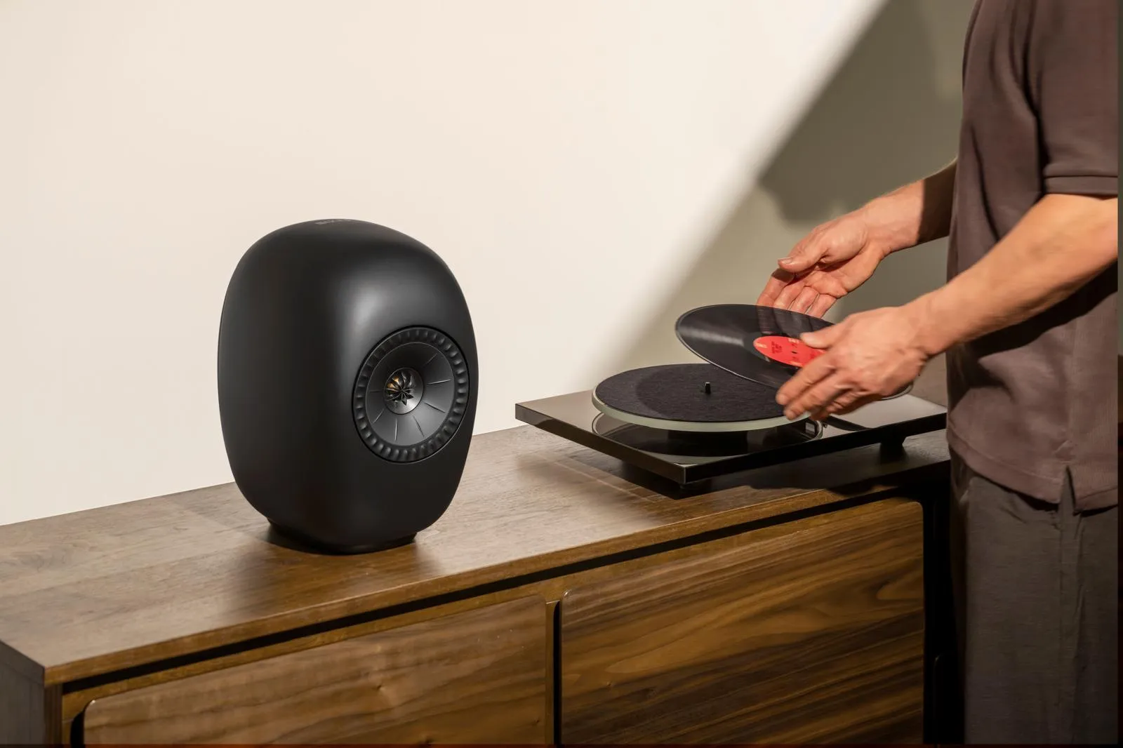 Koyu renk ahşap bir konsolun üzerinde, modern hatlara sahip siyah oval bir hi-fi hoparlörün bitişiğinde, bir kişi elleriyle kırmızı etiketli siyah bir plağı hassasça pikabın tabağına yerleştirerek yüksek kaliteli müzik dinleme keyfine hazırlanıyor.