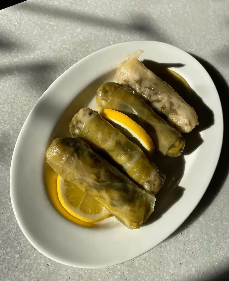Karaköy Lokantası'nın meşhur lezzetlerinden olan beş adet lahana sarması, beyaz oval bir tabakta, keskin ışıkların oluşturduğu uzun gölgeler ve taze limon dilimleri eşliğinde, açık renkli bir yüzeyde özenle sunulmuş şekilde görülüyor.
