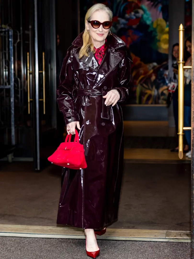Meryl Streep, parlak koyu bordo rugan trençkotu, kırmızı desenli bluzu ve kırmızı sivri burunlu topuklu ayakkabılarıyla, öne çıkan kırmızı Manu Atelier el çantasını taşırken, kedi gözü güneş gözlükleriyle gülümseyerek bir binadan şık bir şekilde ayrılıyor.