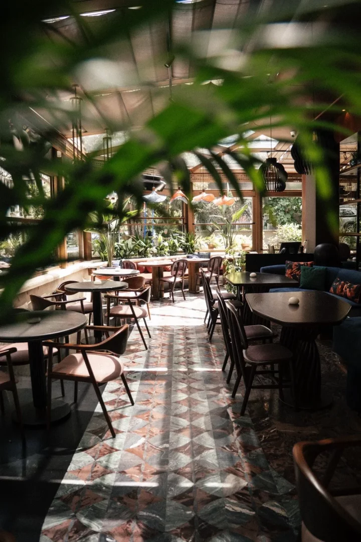 Doğal ışıkla aydınlanmış, bol bitki örtüsüyle çevrili şık bir kafe veya restoranın iç mekanı, desenli mermer zemini ve modern masa sandalye düzenlemeleriyle sıcak bir atmosfer sunarken, ön plandaki yemyeşil yapraklar derinlik ve tazelik katıyor.