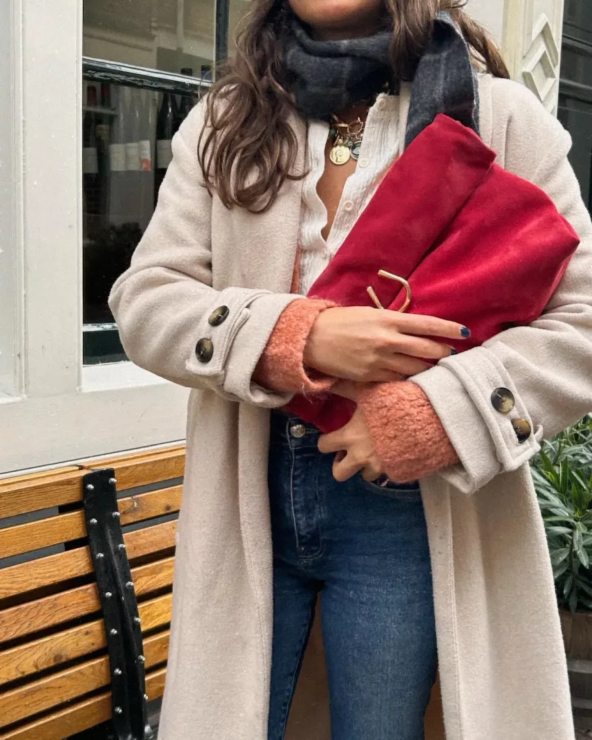 Uzun açık bej bir palto, beyaz bir gömlek, boynunda katmanlı altın kolyeler ve koyu gri bir atkı ile mavi kot pantolon giymiş bir kadın, turuncu kollu kazağı paltonun altından görünen ve tırnaklarında mavi oje bulunan elleriyle altın detaylı parlak kırmızı bir el çantasını tutarken, arka planda ahşap bir bank ve yeşilliklerin olduğu dış mekanda duruyor.
