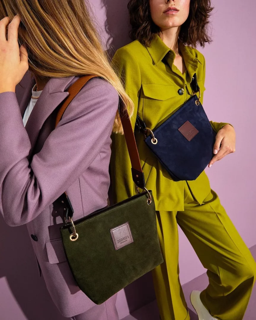 Megna Studio süet çanta koleksiyonundan iki farklı rengi sergileyen bu görselde, mor blazer ceketli bir kadın koyu yeşil bir omuz çantası taşırken, misket yeşili pantolon takımlı diğer bir kadın lacivert bir çanta tutuyor, mor duvara yaslanmış şık duruşlarıyla çantaların dokusunu ve renklerini vurguluyorlar.