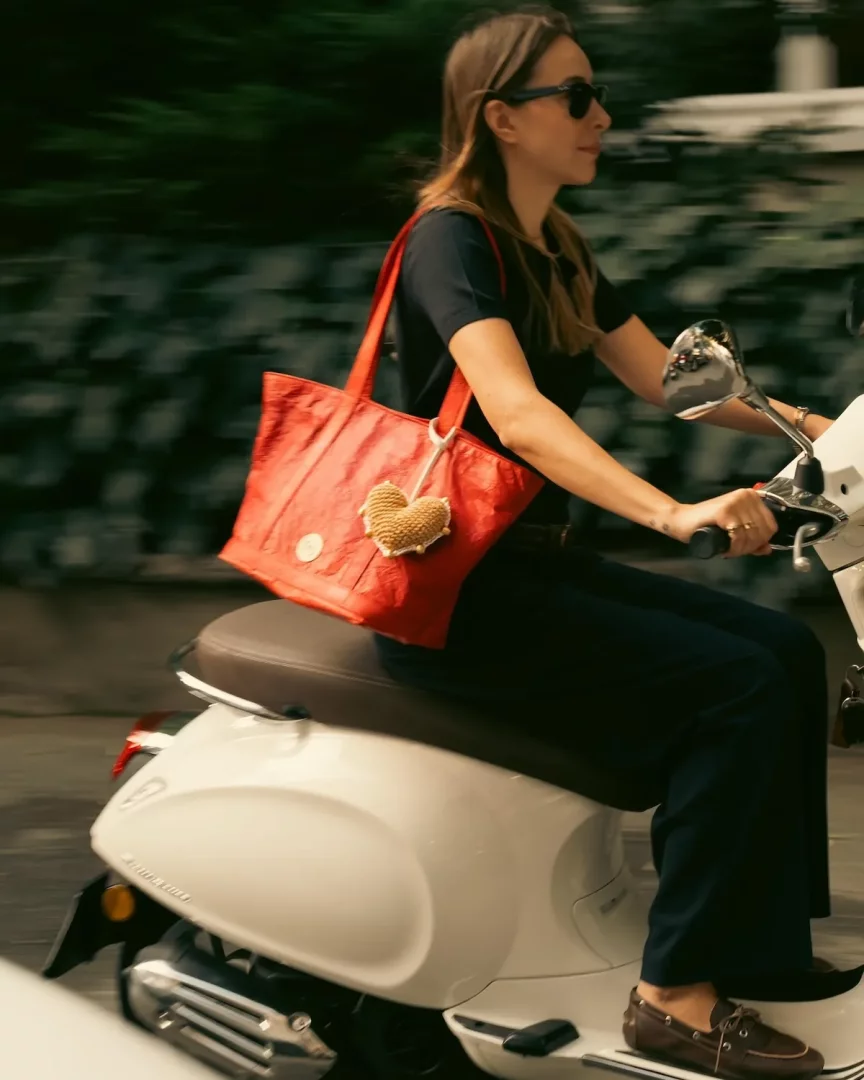 Hızla ilerleyen beyaz bir scooter üzerinde, siyah tişörtü ve koyu renk pantolonuyla güneş gözlüğü takan genç bir kadın, omuzunda örgü kalpli bir anahtarlıkla süslenmiş parlak kırmızı bir Mehry Mu tote çanta taşıyor.