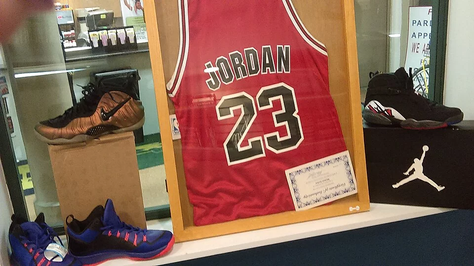 Michael Jordan'ın efsanevi kariyerindeki başarılarını simgeleyen, bir sertifikayla birlikte ahşap çerçeve içinde sergilenen 23 numaralı ikonik kırmızı forması, etrafında farklı modellerde koleksiyonluk spor ayakkabıları ile bir vitrini dolduruyor.