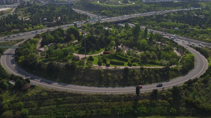Nezahat Gökyiğit Botanik Bahçesi'nin yukarıdan çekilmiş bir görüntüsü, yemyeşil ağaçlar ve düzenli patikalarla dolu dairesel bir alanı sergilerken, bahçeyi saran yoğun trafikli çok şeritli otoyollar ve uzaklardaki binalar, bu eşsiz doğal yaşam alanının şehirle olan bağlantısını vurguluyor.