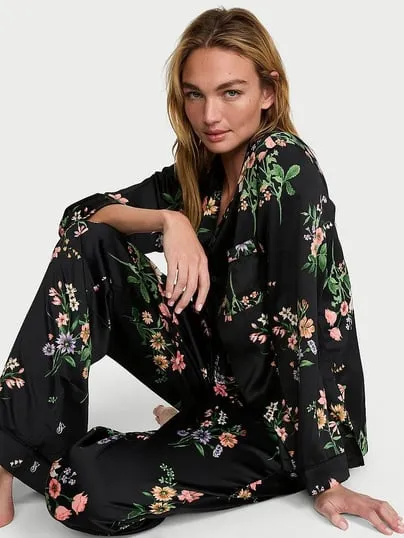Sarışın model, siyah zemin üzerine pembe, turuncu, mor ve yeşil tonlarda canlı çiçek desenleriyle süslenmiş, Victoria's Secret markalı ipeksi bir pijama takımı giymiş, yere oturmuş poz veriyor ve modern pijama şıklığını vurguluyor.