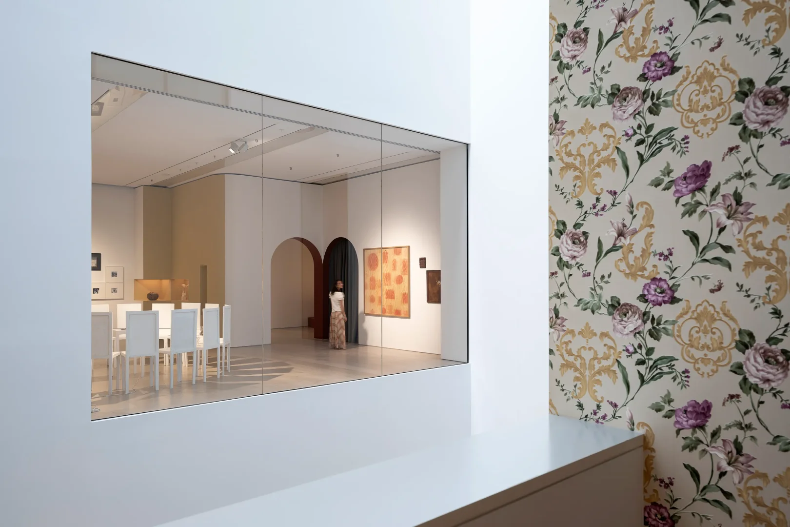 Beyaz duvarlı, kemerli geçitleri olan aydınlık bir çağdaş sanat galerisinin cam pencereden görünen iç kısmında, desenli pantolonlu bir ziyaretçi sergilenen tablolardan birine bakarken, sağda mor güller ve altın barok desenli duvar kâğıdı olan bir duvar ve solda beyaz masalar ile sandalyeler görülüyor.