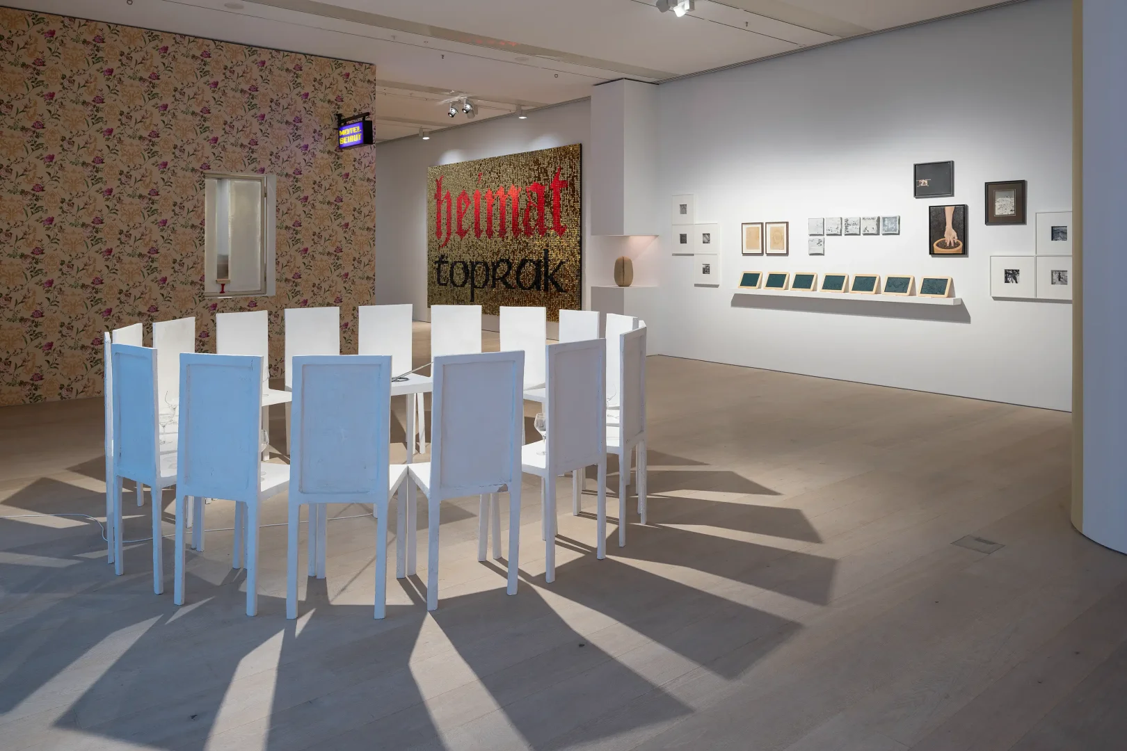 Arter’deki bir çağdaş sanat sergisinden alınan bu karede, aydınlık bir salonda dairesel şekilde düzenlenmiş beyaz sandalyeler ve masanın oluşturduğu yerleştirme, arka planda çiçek desenli duvar kağıdının üzerindeki ayna detayı ve duvardaki heimat toprak yazılı büyük eserle dikkat çekerken, sağ duvarda çeşitli küçük çerçeveli işler sergileniyor.