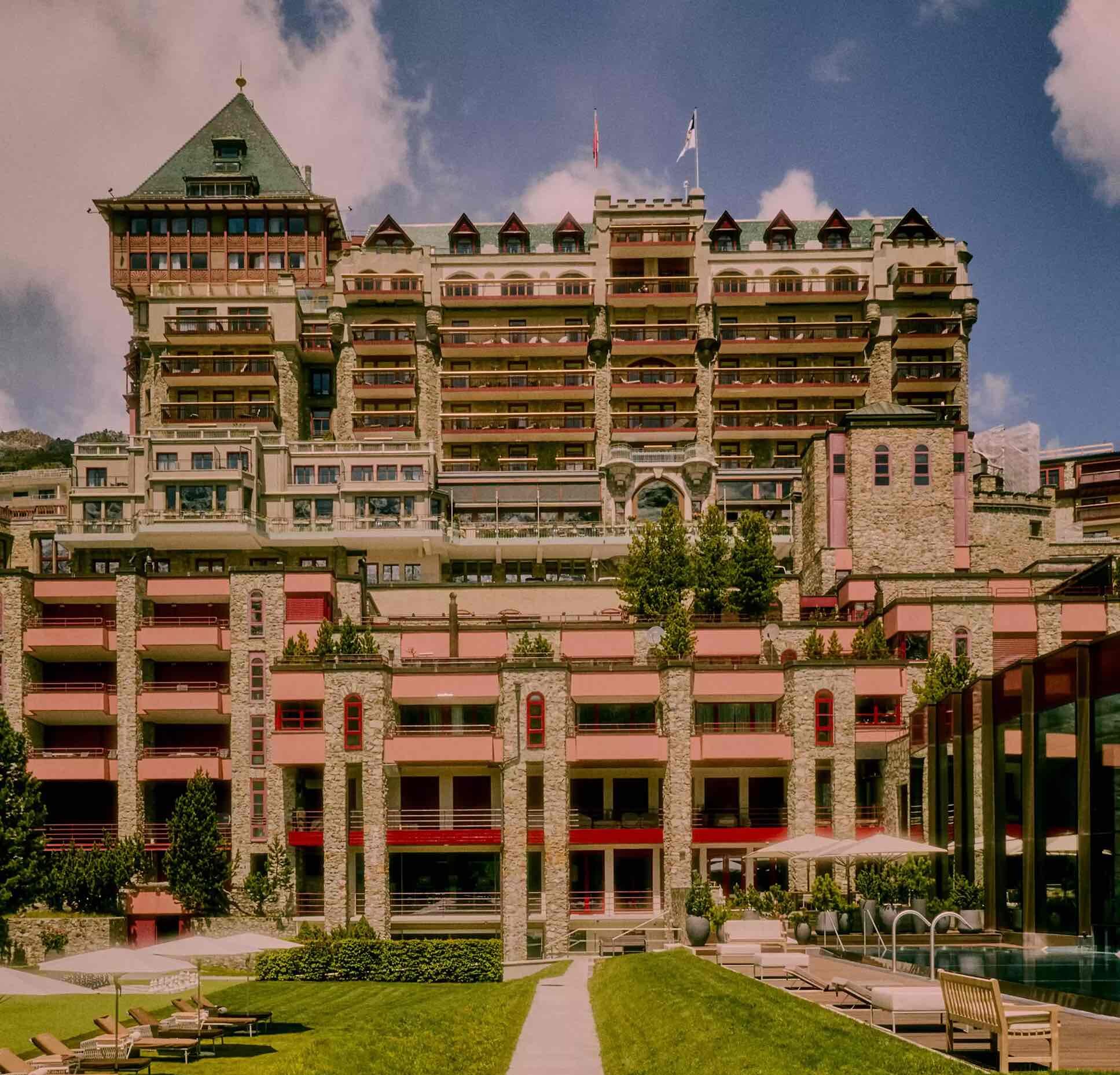 Badrutt's Palace'ın büyüleyici mimarisi, taş cephesi, katmanlı kırmızı tenteli balkonları ve sivri yeşil çatısı ile dikkat çekerken, ön plandaki yemyeşil çim alan, şezlonglar ve berrak bir yüzme havuzu, masmavi gökyüzü altında lüks bir tatil atmosferi oluşturuyor.