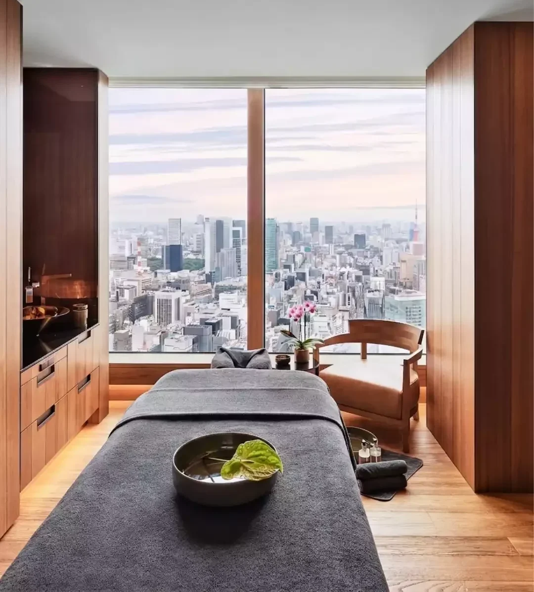 Bulgari Tokyo'nun lüks spa odası, geniş penceresinden Tokyo şehir manzarası sunan, üzerinde koyu gri örtü ve bir kase yaprak bulunan masaj yatağı, ahşap sandalye ve zarif pembe orkidelerle huzurlu bir dinlenme köşesi oluşturuyor.