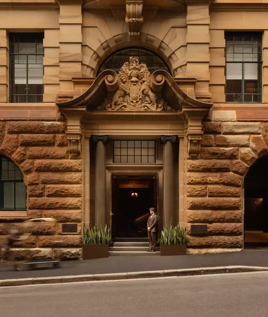 Capella Sydney otelinin bej taş bloklardan inşa edilmiş, oymalı bir amblemle taçlandırılmış kemerli girişinde, üniformalı bir doorman kapı önündeki merdivenlerin başında beklerken, yanlarda bitki saksıları ve sol tarafta hareketli bir figür görülüyor.
