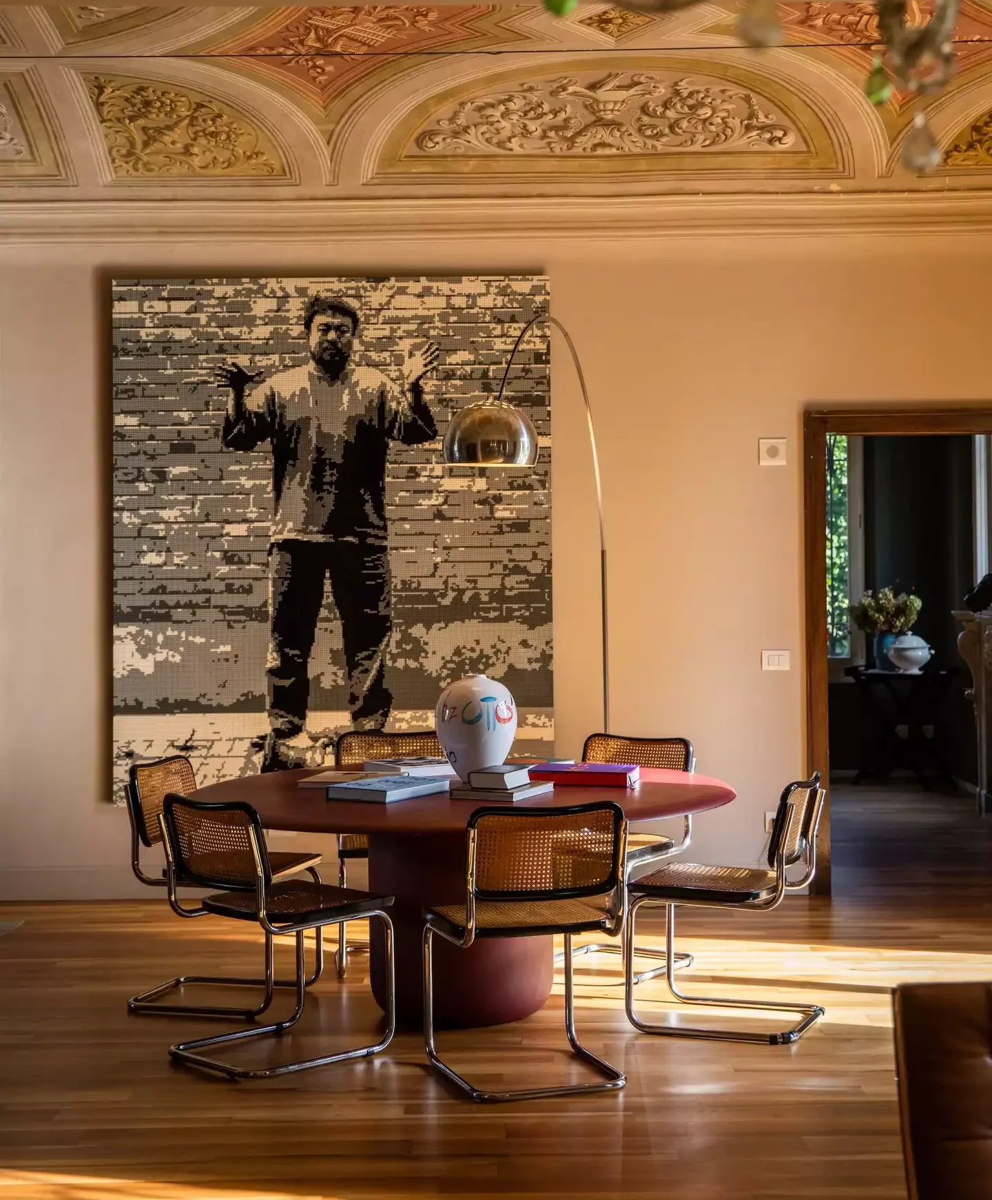 Casa Maria Luigia'nın zarif iç mekanında, geleneksel kemerli freskli tavanın altında, elleri havada, tuğla desenli bir arka plan önünde duran bir adamı tasvir eden modern bir piksel sanat eseri dikkat çekerken, bu dev eserin önünde kitaplar ve sanatsal bir vazo ile süslenmiş yuvarlak bir masa etrafına dizilmiş rattan sandalyeler ve ahşap zemin üzerine düşen gün ışığı, eşsiz bir ambiyans sunuyor.