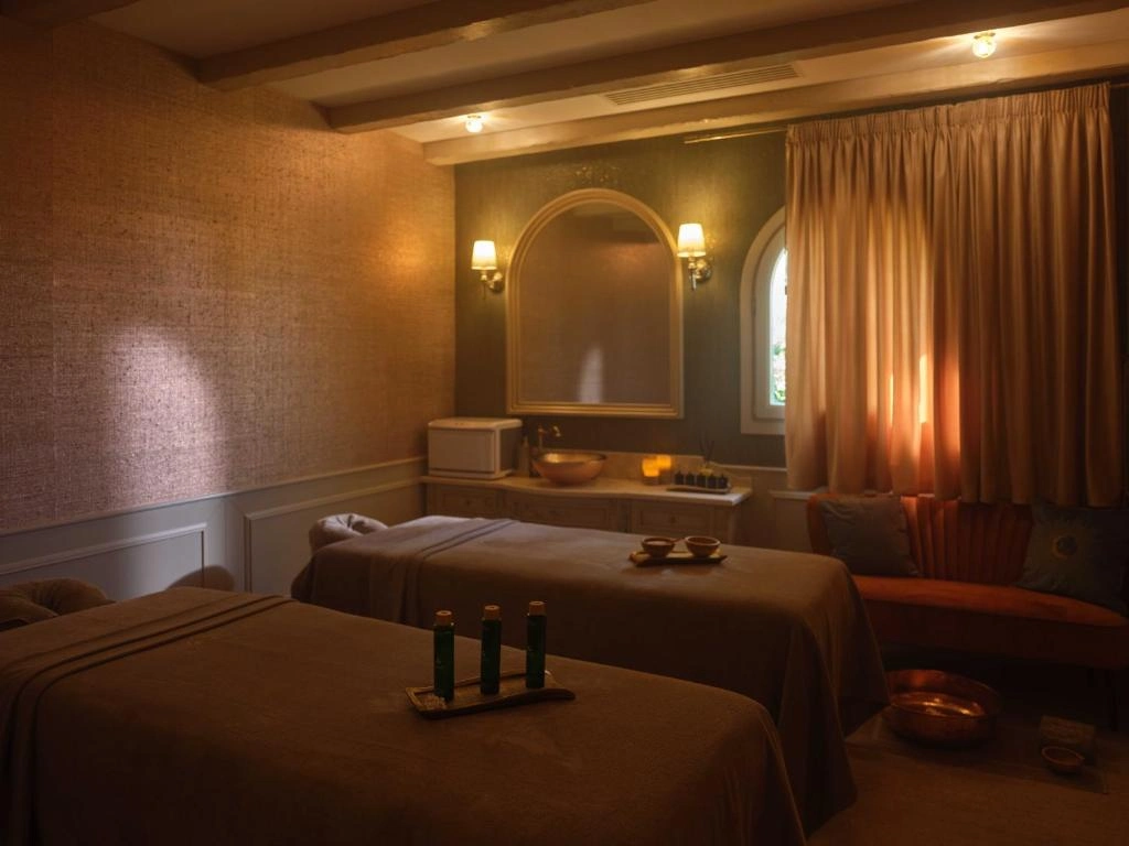 Loş ışıklandırılmış, sıcak tonlarda dekore edilmiş lüks bir spa odasında, üzerinde yeşil şişeler ve küçük kaseler bulunan iki masaj masası, duvar aynası, kadife kanepe ve bakır detaylar rahatlatıcı bir ortam sunuyor.