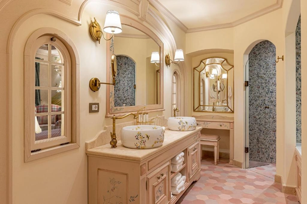 Casa Timis'teki bu şık banyoda, çiçek desenli iki lavabo, mermer tezgah ve altın armatürlerle bezeli çift makyaj ünitesi, kemerli pencereden yatak odası görünümü, desenli duvar kağıtlı duş alanı ve pastel tonlarda altıgen yer karoları lüks bir konaklama deneyimi sunuyor.