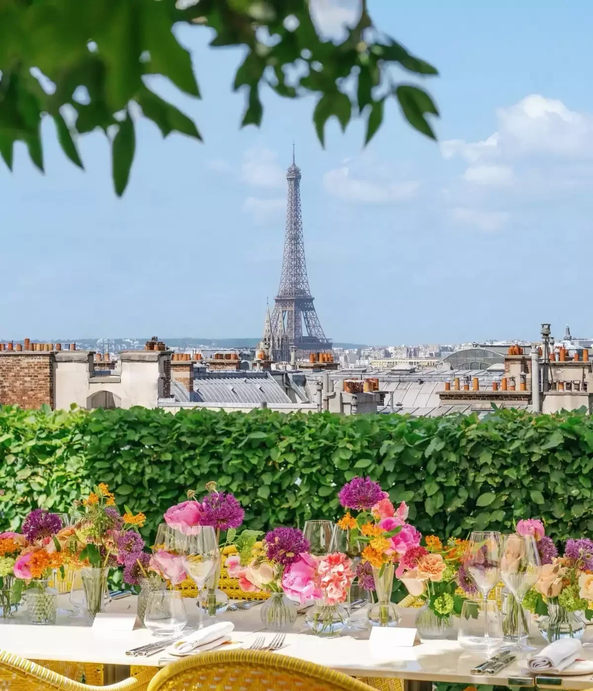 Paris'in çatılarında, Eyfel Kulesi manzaralı bir terasta, canlı renklerde çiçek aranjmanları ve şık masa düzenlemeleriyle bezeli uzun bir yemek masası keyifli bir açık hava yemeği için hazırlanmış.