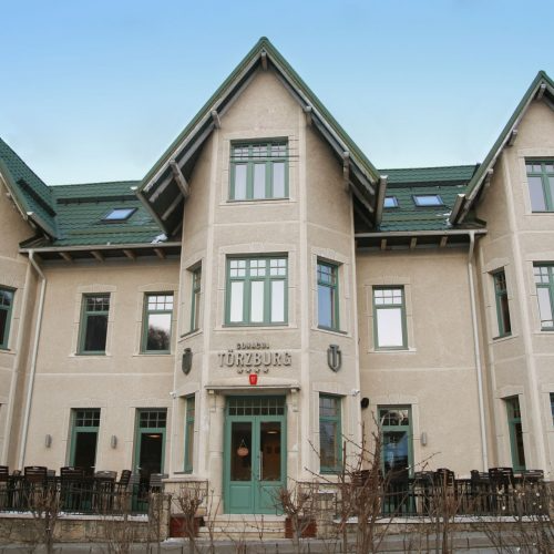 Hotel Türzburg yazılı tabelası bulunan, sivri çatıları, koyu yeşil çerçeveli pencereleri ve bej renkli cephesiyle dikkat çeken, zarif ve tarihi bir konak veya otel binasının dış görünüşü, önündeki terasta masa ve sandalyelerin de yer aldığı geniş açıyla, parlak bir gökyüzü altında gösterilmektedir.