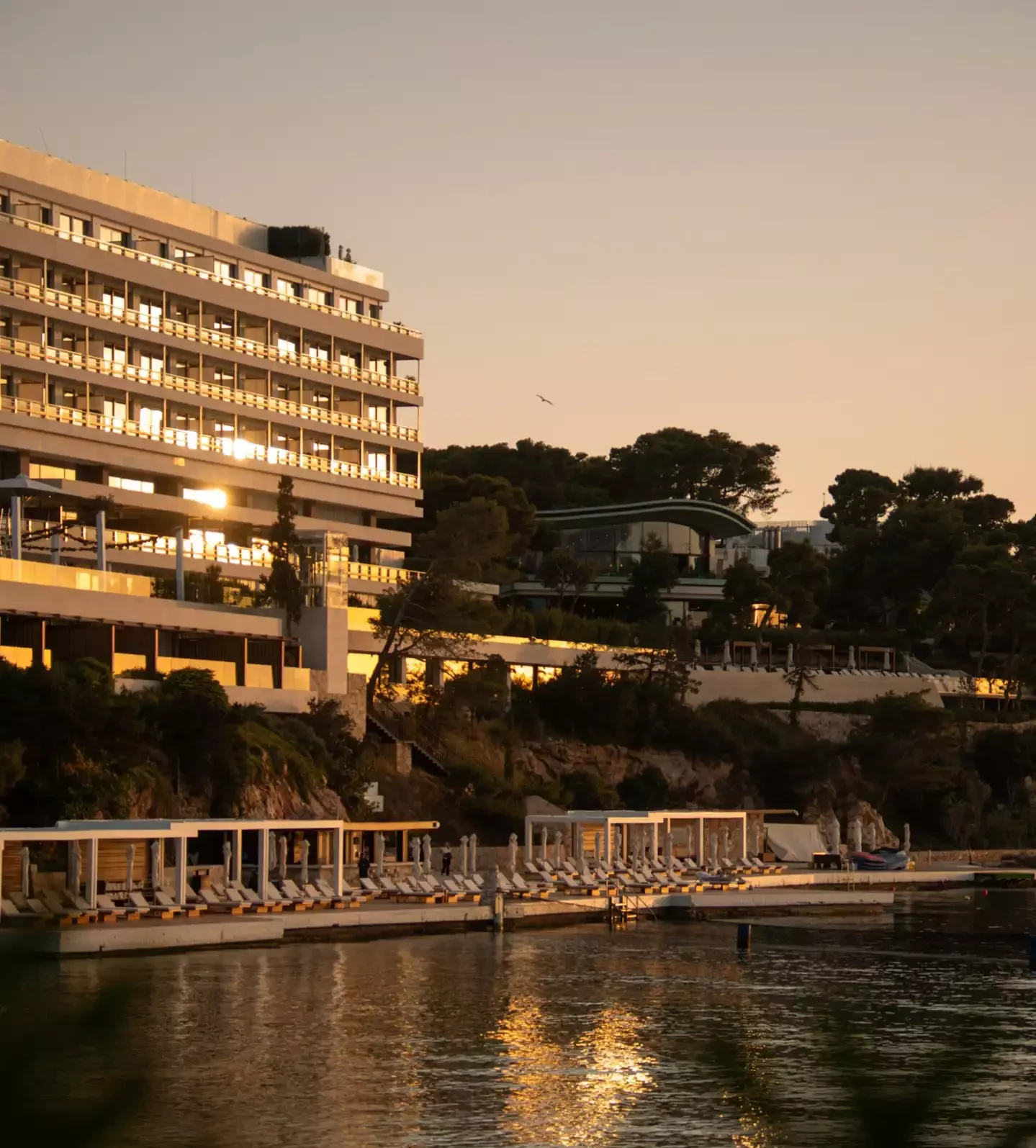 Gün batımının altın rengi ışıkları altında, Four Seasons Astir Palace otelinin camları ve önündeki denizin yüzeyi parıldarken, sahildeki şezlonglu lüks dinlenme alanları ile çevrili yemyeşil tepelerin eteklerindeki bu görkemli yapı, huzurlu bir Akdeniz atmosferi sunuyor.