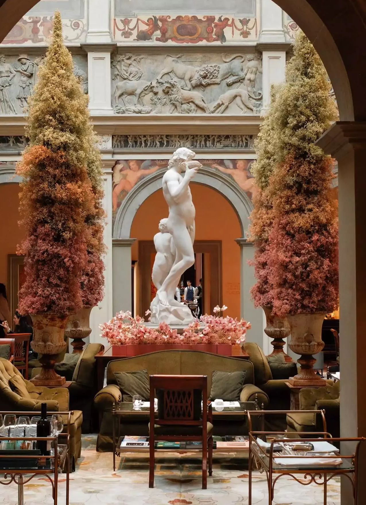 Four Seasons Firenze'deki görkemli lobide, iki katlı salonun merkezinde mermer bir Bacchus heykeli ve yanında duran küçük figür, sonbahar renklerinde devasa çiçek aranjmanları ile çevrili. Arkada detaylı freskler ve kabartmalar, ön planda ise konforlu yeşil kadife oturma grupları ve zarif masalar, bu lüks mekanı tamamlıyor.
