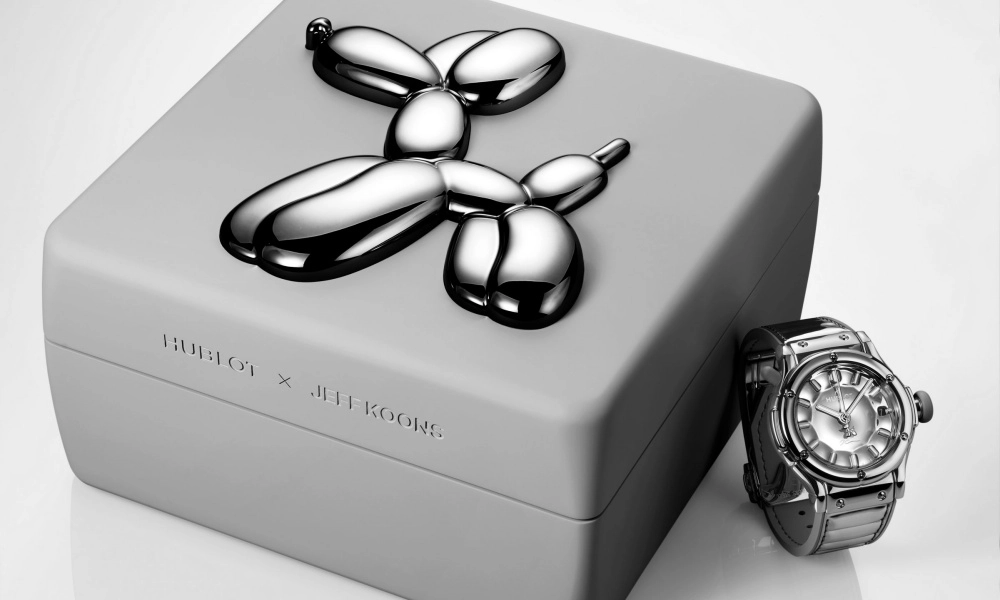 Açık gri bir kutu üzerinde parlayan, gümüş renkli Jeff Koons imzalı bir balon köpek heykelciği ile yanında metalik kasalı, gümüş kadranlı bir Hublot Classic Fusion saatin görüldüğü, sanat ve saatçiliğin buluştuğu bir kompozisyon.