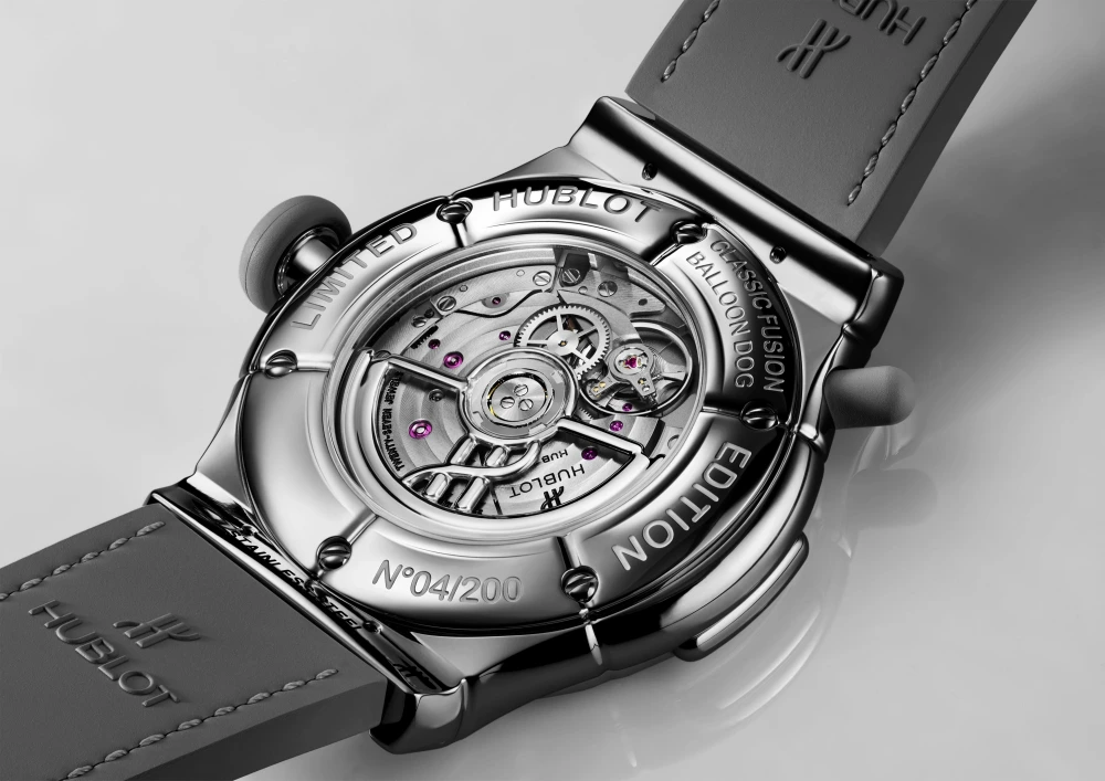 Hublot Classic Fusion Balloon Dog Limited Edition saatinin arkadan görünümü, karmaşık gümüş mekanizmasını ve pembe rubin taşlarını şeffaf bir pencereden gözler önüne sererken, gümüş renkli kasasında N°04/200 seri numarası ve gri kauçuk kayışı dikkat çekiyor.