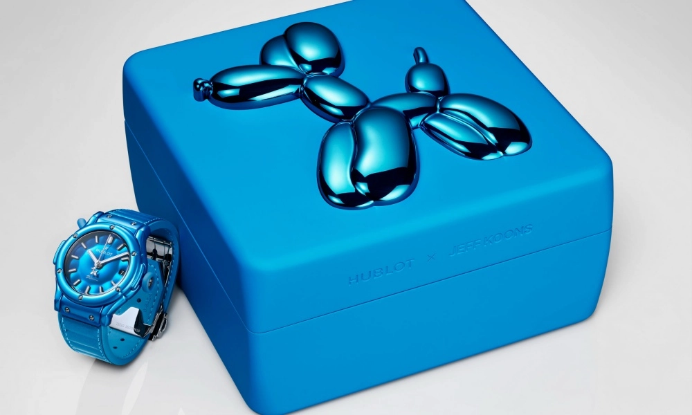 Açık gri zemin üzerinde konumlanmış, dikkat çekici mavi kadran ve kordona sahip bir Hublot Classic Fusion saati, sağında ise sanatçı Jeff Koons'un ikonik parlak mavi balon köpek heykeliyle süslenmiş bir kutu sergileniyor.