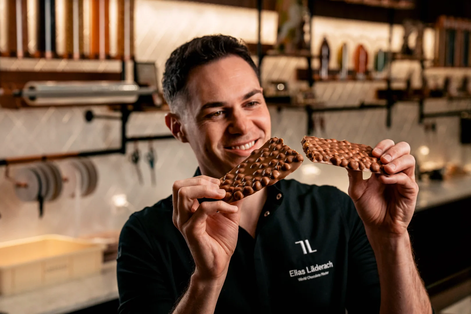 World Chocolate Master unvanlı Elias Läderach, elinde FrischSchoggi adı verilen, taze kırılmış, üzerinde bolca fındık bulunan bir sütlü çikolata parçasını gülümseyerek inceliyor; bu an, iyi çikolatanın zanaat ve tazelikle buluştuğunu gösteriyor.