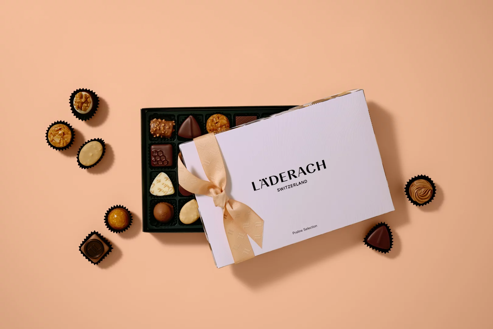 LÄDERACH SWITZERLAND yazılı, kurdeleli beyaz kutunun içinden ve etrafına dağılmış, farklı dolgu ve görünümlere sahip çikolatalı pralinler, markanın iyi çikolata anlayışını, zanaatını ve tazeliğini yansıtan davetkar bir seçki sunuyor.