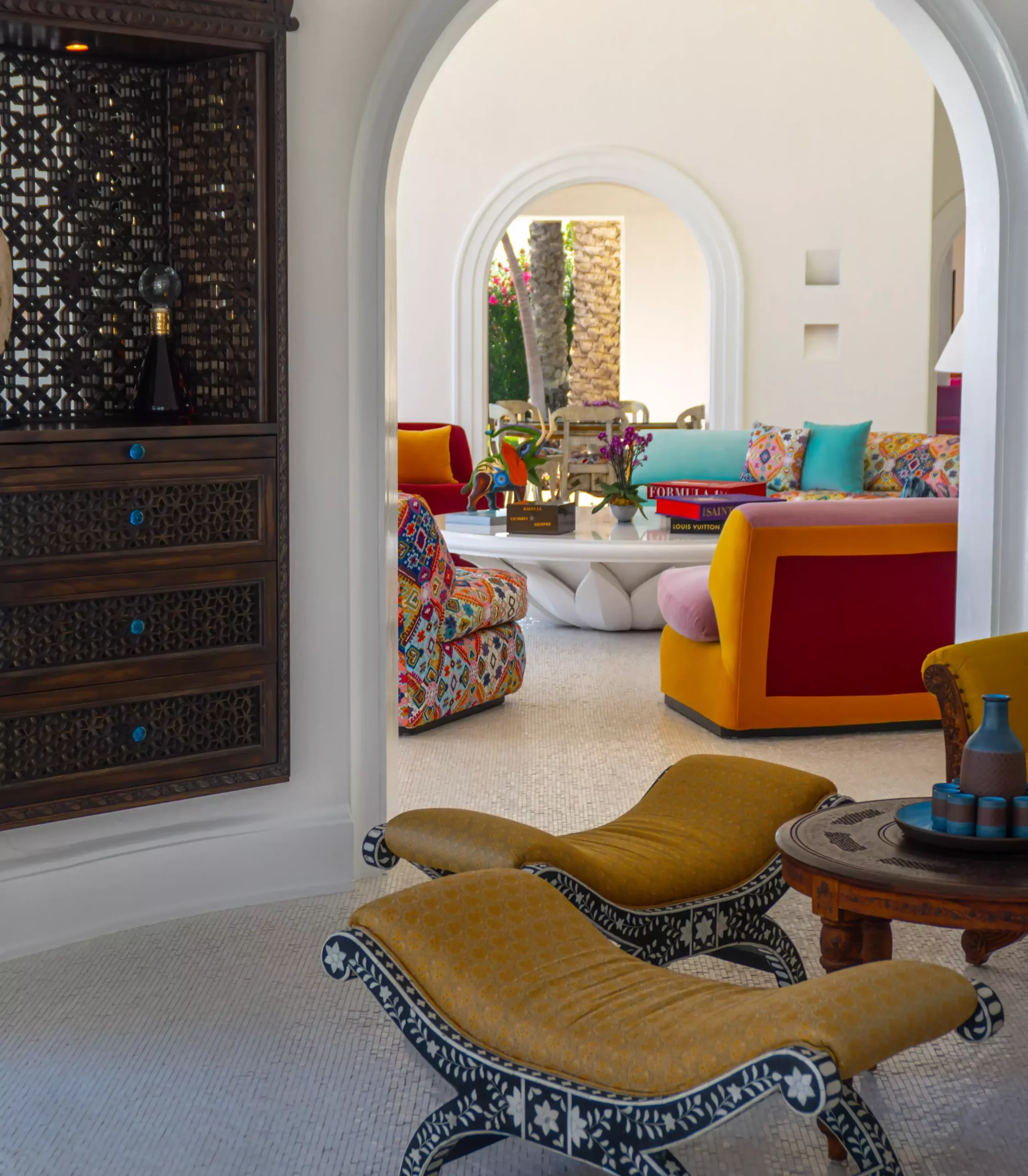 Las Ventanas al Paraíso'daki lüks bir yaşam alanının detaylarını gösteren bu görüntüde, ön planda zarif oymalı koyu ahşap iskeletlere ve sarı döşemelere sahip iki şezlong ile arkada kemerli bir kapıdan görünen canlı renkli mobilyalar ve tropikal bahçe manzarası cennet gibi bir atmosfer sunuyor.