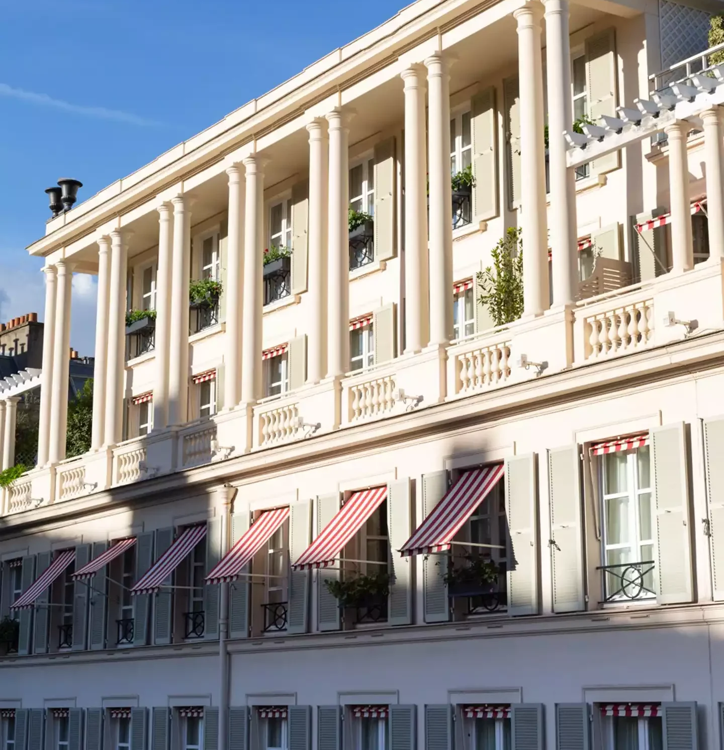 Le Bristol otelinin klasik mimariye sahip bej rengi dış cephesi, sütunlu üst katları, kırmızı-beyaz çizgili tentelerle süslenmiş pencereleri ve dekoratif panjurlarıyla, saksılardaki yeşil bitkilerin güneş ışığında parladığı zarif bir görünüm sergiliyor.