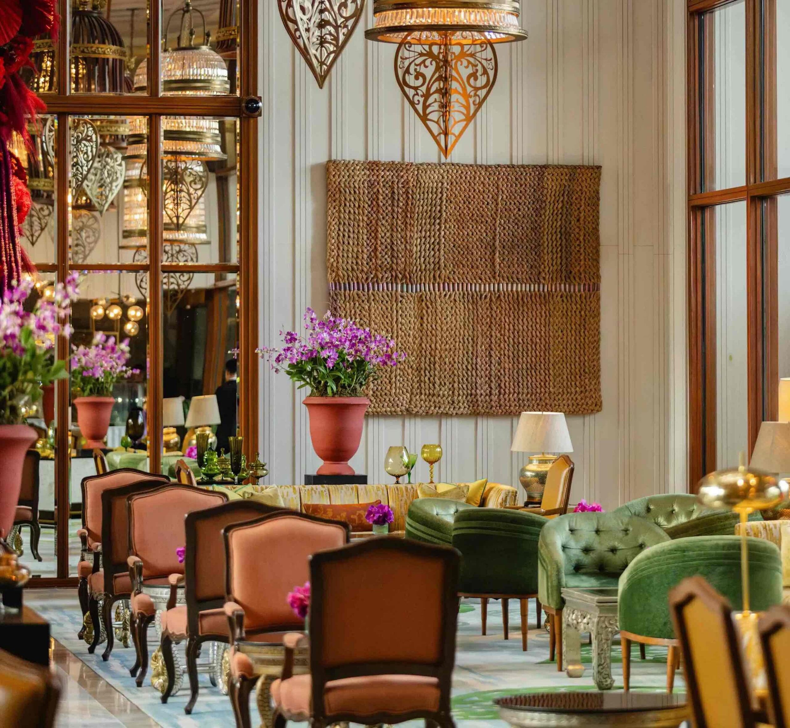 Mandarin Oriental Bangkok gibi lüks bir otel lobisinde, mor orkidelerle dolu büyük bir saksının önünde sıralanmış kahverengi ve yeşil kadife koltuklar, aynalı paneller, hasır dokulu duvar sanatı ve zarif aydınlatmalarla donatılmış göz alıcı ve konforlu bir iç mekan tasarımı görülmektedir.