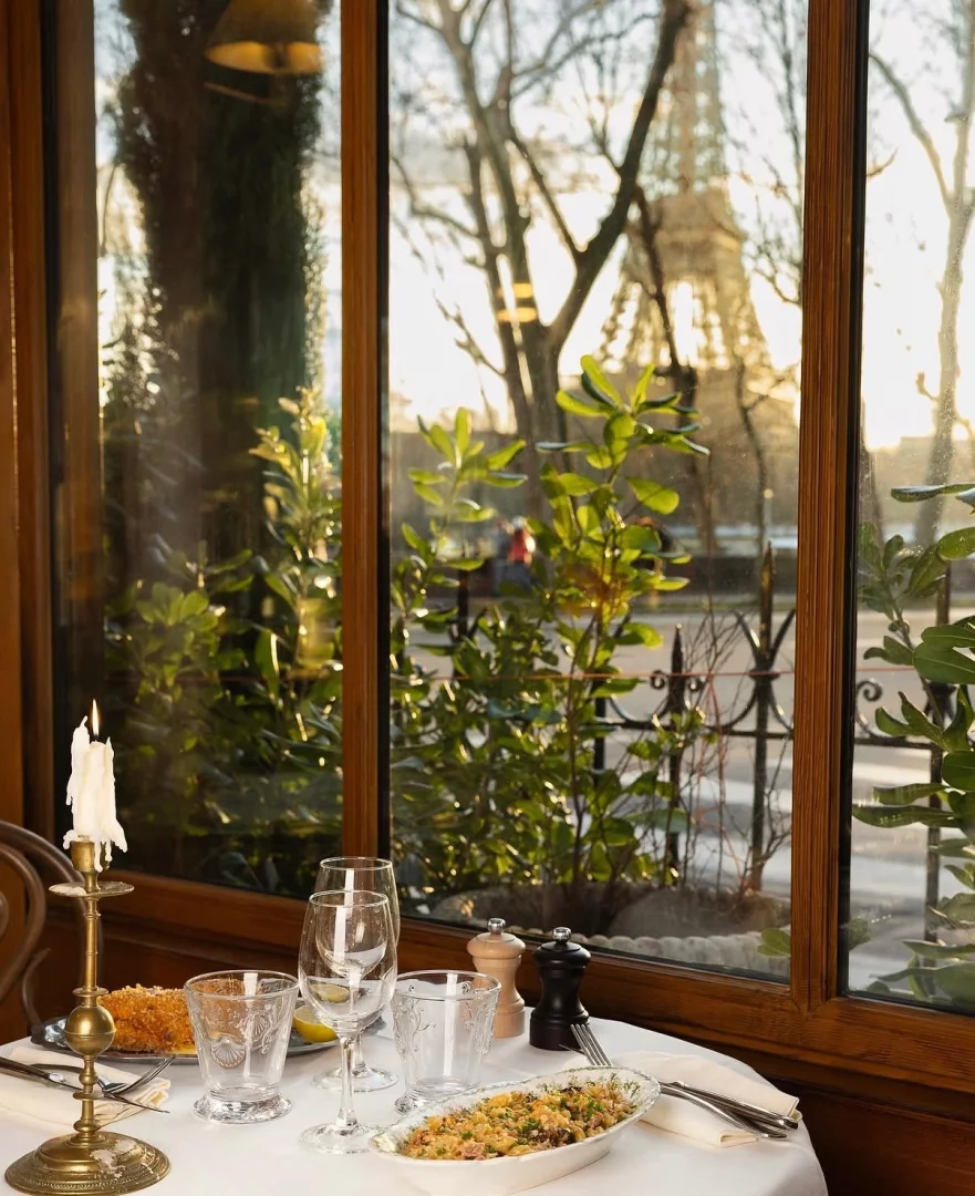 Beyaz masa örtüsü üzerinde yanan bir mum, boş şarap ve su kadehleri ile çeşitli yemeklerin sunulduğu zarif bir restoran masasının önünden bakıldığında, Paris'in akşam güneşiyle altın rengine bürünmüş çıplak ağaçları ve yemyeşil bitkileri arasından Eyfel Kulesi'nin görkemli silüeti pencereden izleniyor.