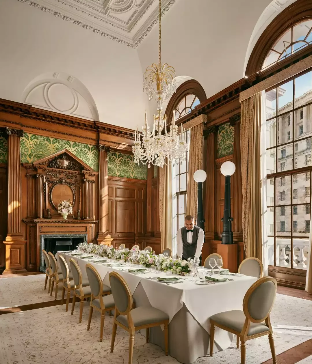 Raffles London at The OWO'daki zarif yemek salonunda, özenle hazırlanmış uzun yemek masası çiçek aranjmanları ve parlak sofra takımlarıyla donatılmış, garsonun konukları ağırlamak için son dokunuşları yaptığı lüks bir ziyafet ortamı sergileniyor.