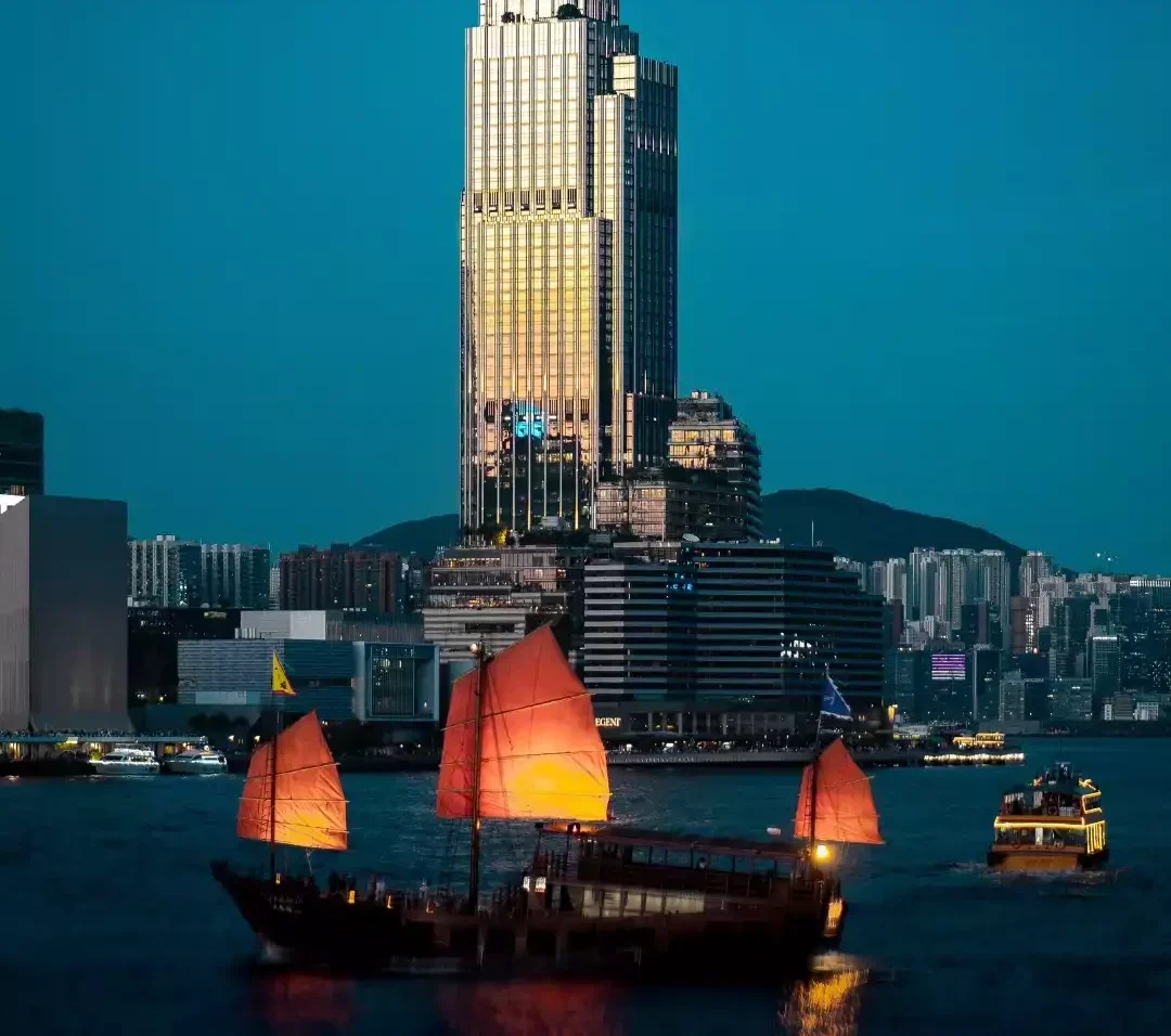 Hong Kong silüetinde alacakaranlıkta parıldayan Rosewood Hong Kong otelinin önünde, üç parlak turuncu yelkeni içten aydınlatılmış geleneksel bir junk teknesi ve diğer tekneler denizde süzülürken, şehrin ışıltısı suya yansıyor.