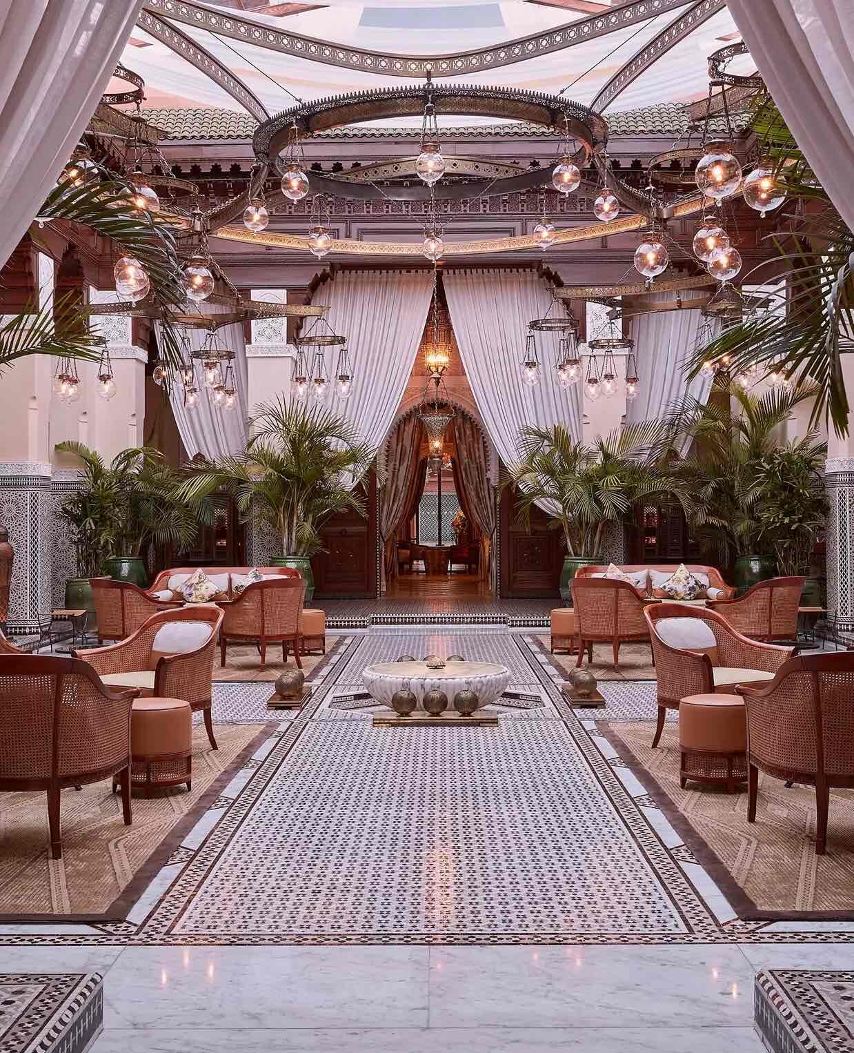 Bir otel lobisi veya lüks bir salona benzeyen bu gösterişli Fas tarzı iç mekanda, ortada uzanan karmaşık desenli mozaik fayanslı bir yol ve yol boyunca karşılıklı yerleştirilmiş hasır koltuk takımları görülüyor; yüksek tavanlardan sarkan zarif küre avizeler ve yeşil palmiye bitkileri, merkezi taş havzanın etrafını sararak mekana ferah ve davetkar bir atmosfer katıyor.