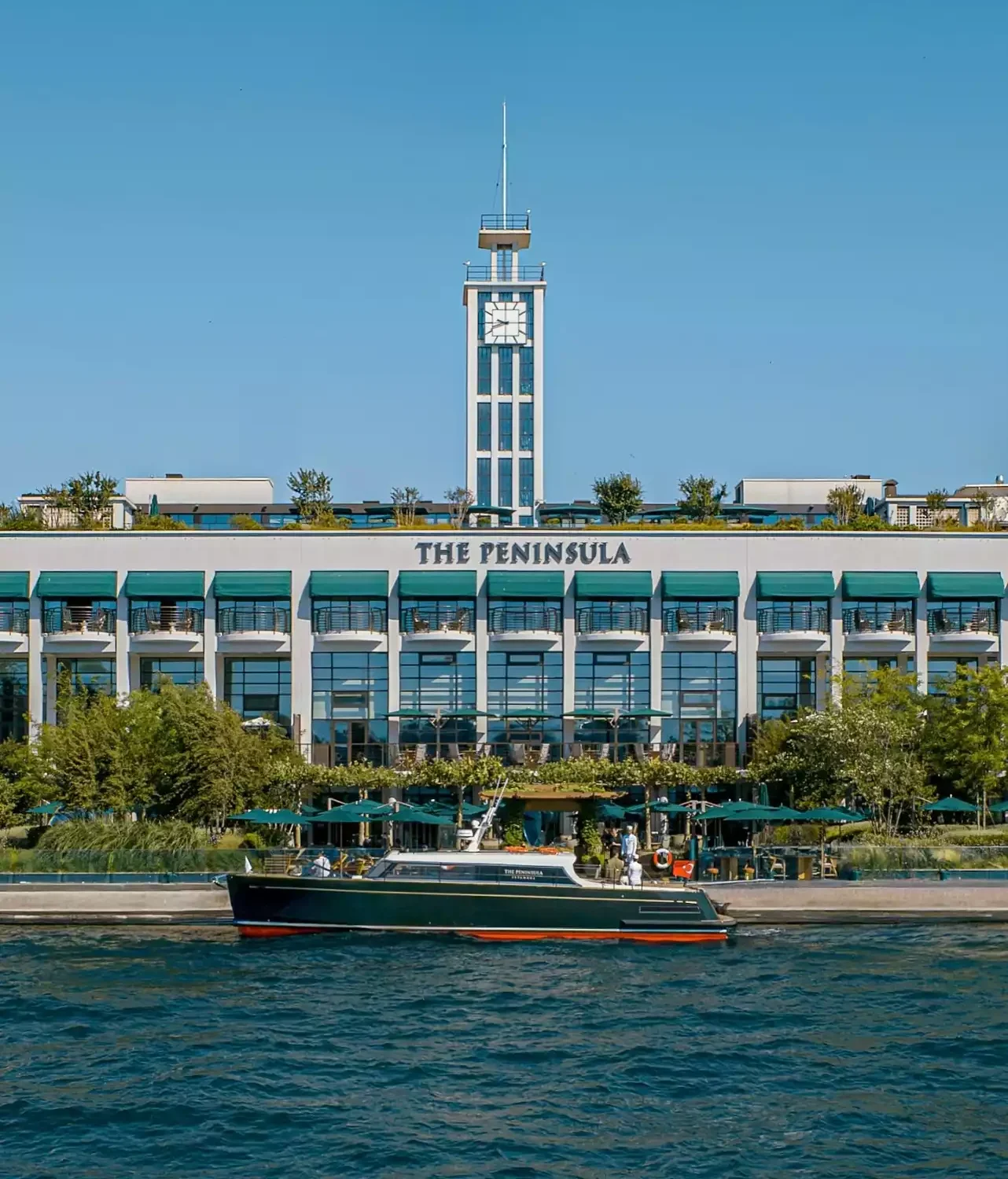 Masmavi bir gökyüzünün altında, ikonik saat kulesiyle dikkat çeken The Peninsula Istanbul oteli, balkonlu ve yeşil tenteli zarif cephesiyle ön plana çıkarken, önündeki sakin denizde The Peninsula logosuna sahip koyu yeşil bir tekne duruyor.