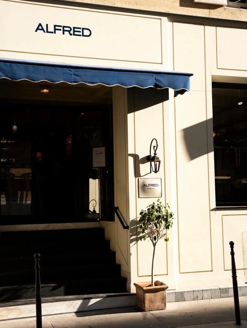 Güneşin aydınlattığı bej renkli Alfred Restaurant'ın dış cephesi, üzerinde belirgin bir şekilde ALFRED yazan bir tabela, altında şık bir mavi tente ve restoranın girişini gösteren merdivenlerin yanında küçük, yeşil yapraklı bir ağaç ile zarif bir fener detayını barındırıyor.