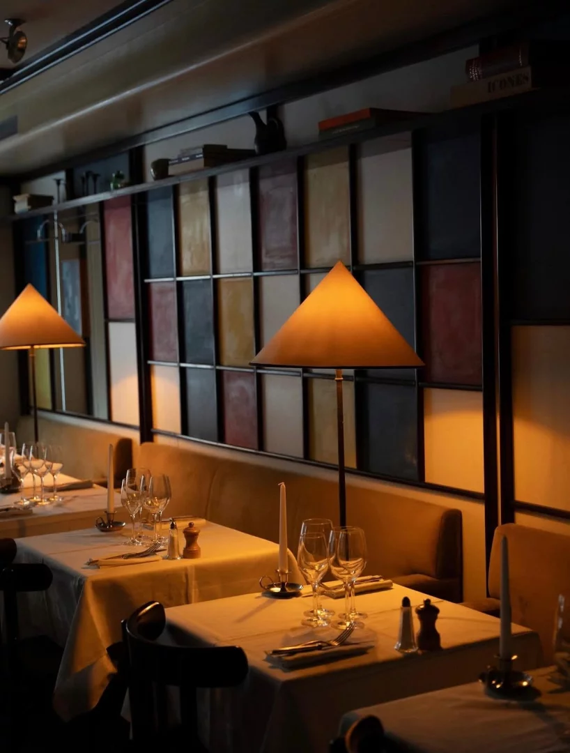 Loş ışıklı bir restoranın sıcak ve davetkar iç mekanında, beyaz örtülü masalar kadeh, çatal-bıçak takımı ve mumlarla özenle düzenlenmişken, üçgen başlıklı ambiyans lambaları özel bir atmosfer yaratıyor; arkadaki renkli kare panellerle bezenmiş duvar ve üzerindeki raf detayları mekanın şık tasarımını tamamlıyor.
