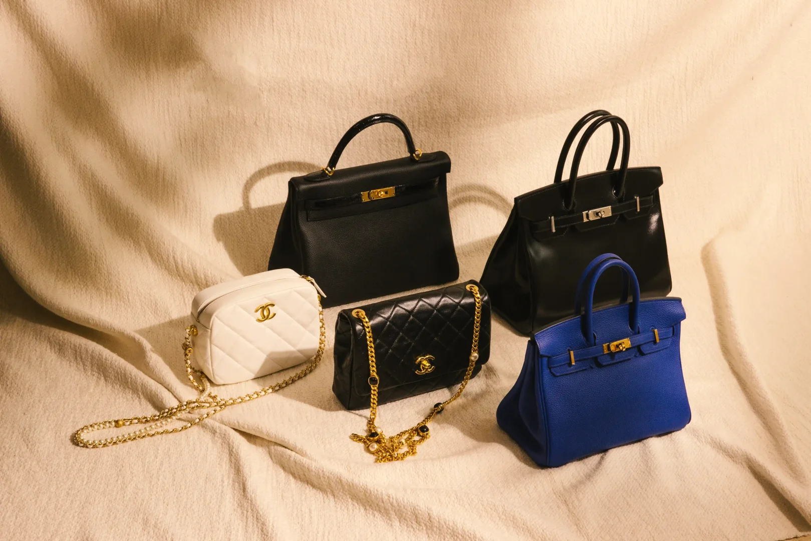 Açık renkli, dokulu bir zemin üzerinde, koleksiyonluk değeri olan beş farklı lüks çanta zarifçe sergileniyor: Hermes Birkin çantalarından biri lacivert, diğeri parlak siyah renkte, bir siyah Hermes Kelly çanta ve altın zincirli, kapitone desenli beyaz ile siyah iki farklı Chanel çanta altın ve gümüş detaylarıyla öne çıkıyor.