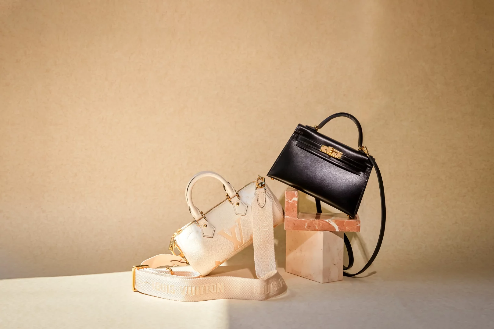 Açık bej tonlarındaki zeminde, sol tarafta logolu geniş kayışıyla krem rengi küçük bir Louis Vuitton çanta, sağ tarafta ise altın detaylı kilidi ve sapıyla siyah renkli, klasik tarzda zarif bir lüks çanta, kahverengi mermer bloklar üzerinde şık bir şekilde sergileniyor.