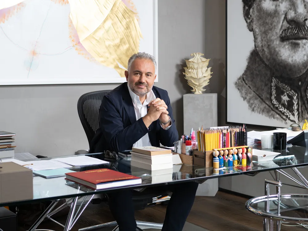 Gri saçlı ve sakallı, koyu renk takım elbiseli bir adam, ellerini kenetlemiş şekilde kameraya bakarak, arka planda modern ve klasik sanat eserlerinin, masasında ise kitaplar ve renkli kalemlerin bulunduğu şık ofisinde, İstanbul'dan bir sanat koleksiyoneri olarak poz veriyor.