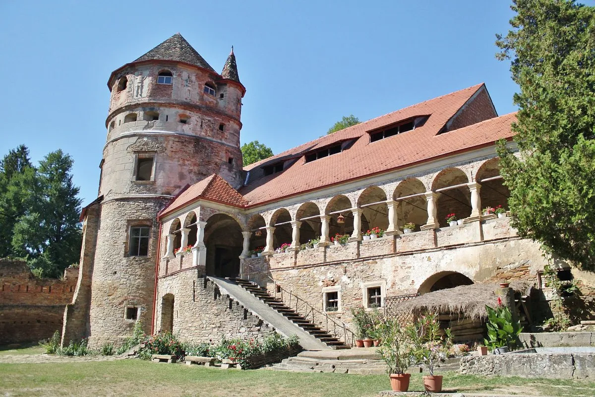 Bethlen Kalesi'nin bu panoramik görüntüsünde, sol tarafta sivri konik çatılı yüksek, yıpranmış taş bir kule, sağ tarafta ise saksı çiçekleriyle bezeli, kemerli bir galeriye ve kırmızı kiremitli bir çatıya sahip uzun bir bina, ön planda basamaklı bir giriş ve çevresi ağaçlarla çevrili yeşil bir alanla birlikte masmavi gökyüzünün altında parlıyor.